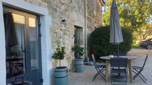 En terrasse med et bord og stole, grill, plantekrukker og en bil i baggrunden.