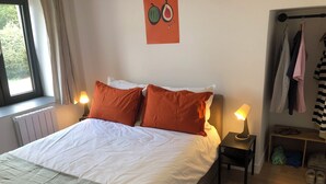 En seng med hvidt og orange sengetøj, to sengeborde med lamper, en radiator, et vindue med gardiner, et billede af frugt på væggen, og et tøjstativ med forskelligt tøj.