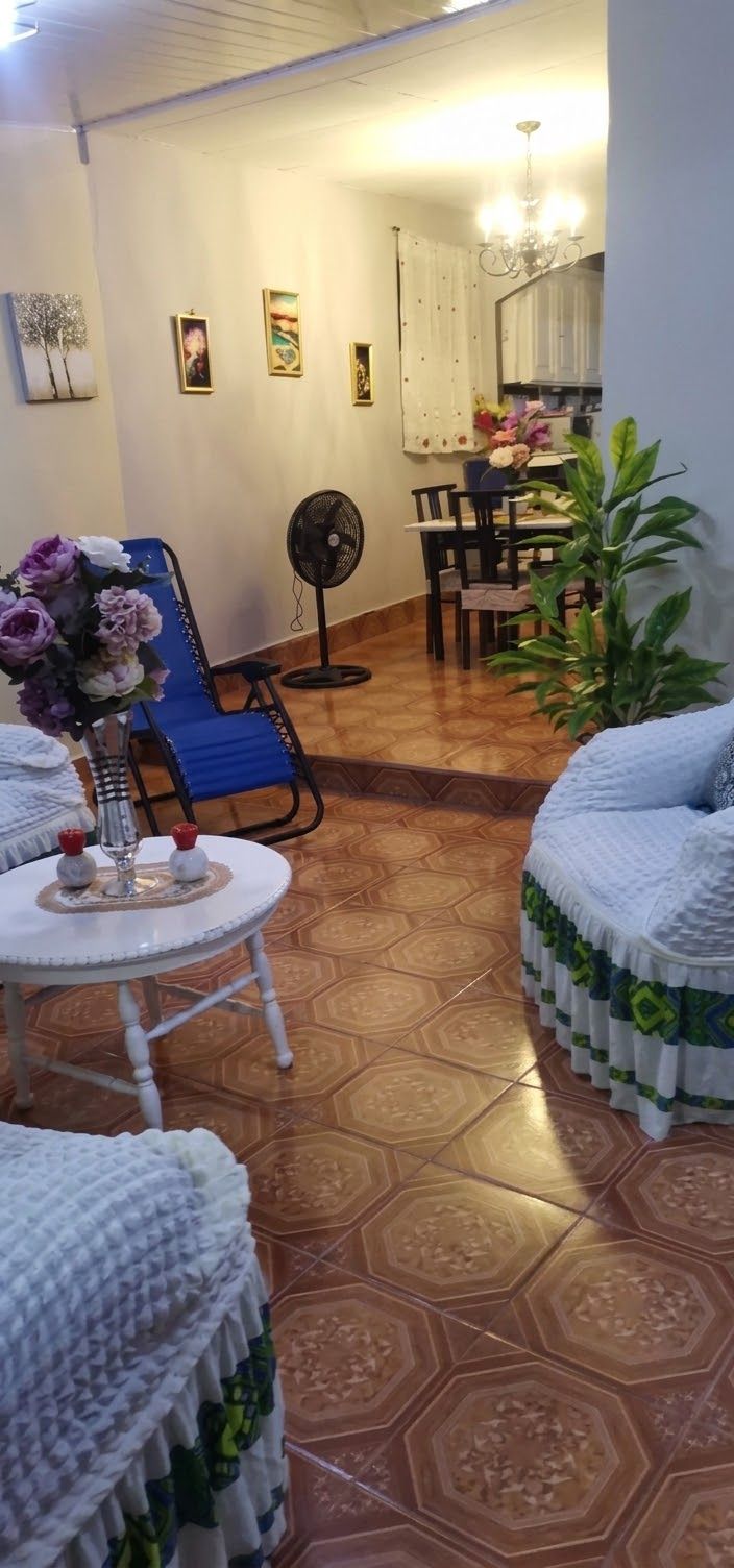 A living room with a blue armchair, a round table with a vase of flowers, a fan, and a dining area with chairs and a table.