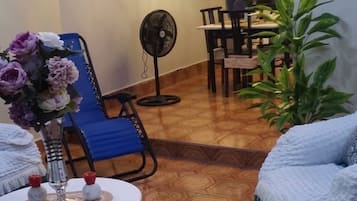 A living room with a blue armchair, a round table with a vase of flowers, a fan, and a dining area with chairs and a table.
