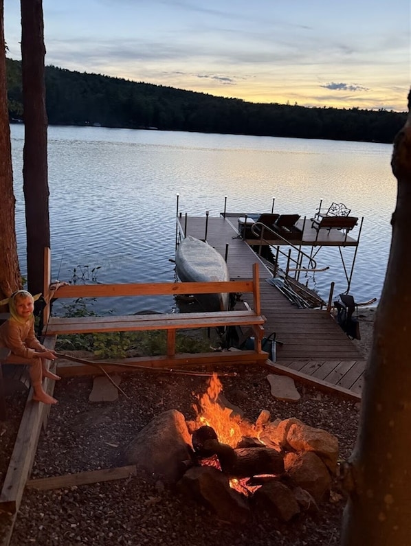 A wooden dock with a bench and a fire pit, overlooking a calm lake with a dock in the background.