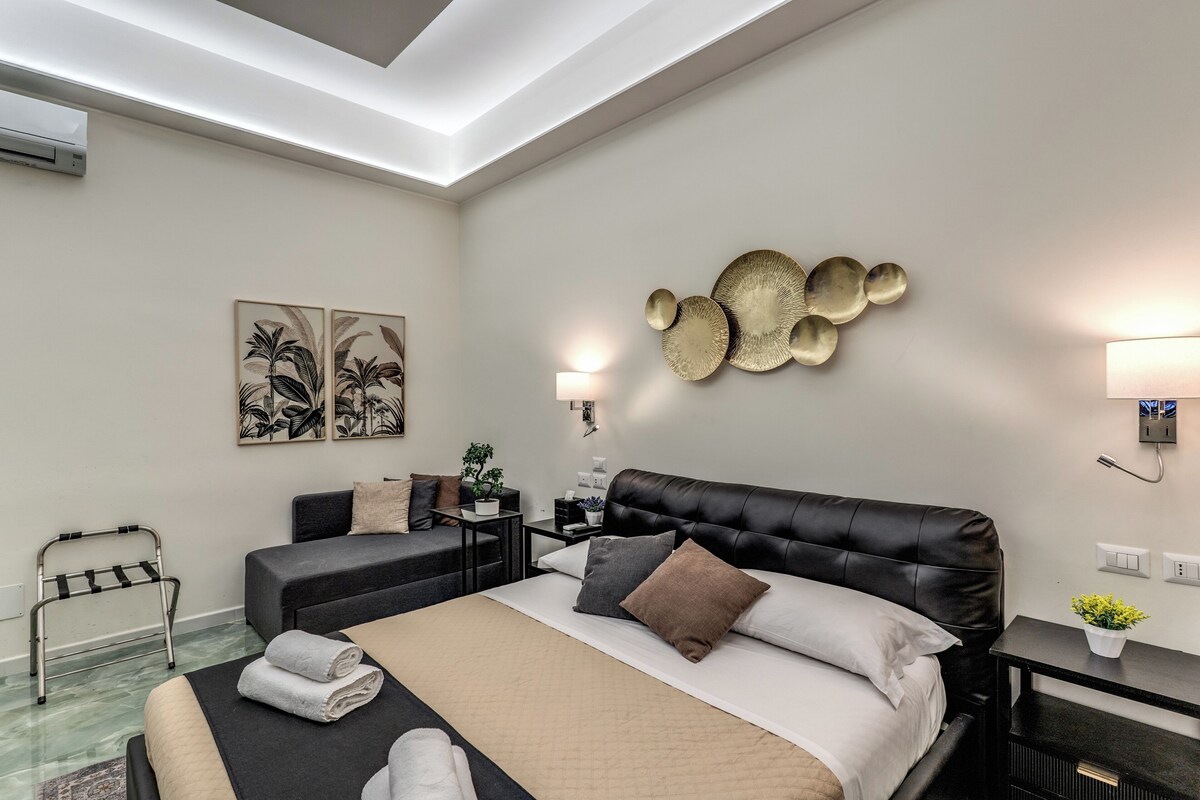 A modern hotel room with a large bed, bedside tables, a small sofa, a chair, and wall art.