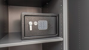 A safe with a keypad and a keyhole, placed on a shelf in a room with a plain wall and a metal frame. - Adriana 5 Luxury rooms (Rome)