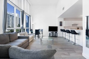 Ein modernes Wohnzimmer mit einer Couch, Sesseln, einem Glastisch, einem Fernseher und einer Küchenzeile mit Barhocker.