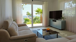 A living room with a sofa, a coffee table, a TV on a stand, and a view of the outdoors. - Oasis Villa - Villa with pool and private giarino, San Remo (Sanremo)