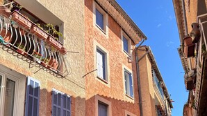 Une rue étroite bordée d’immeubles dotés de balcons ornés de plantes en pot, d’une porte bleue et d’un mur en pierre.