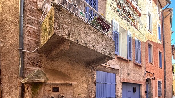 Une rue étroite avec une fontaine en pierre, une rambarde en métal et un bâtiment doté d’une porte et de volets bleus.