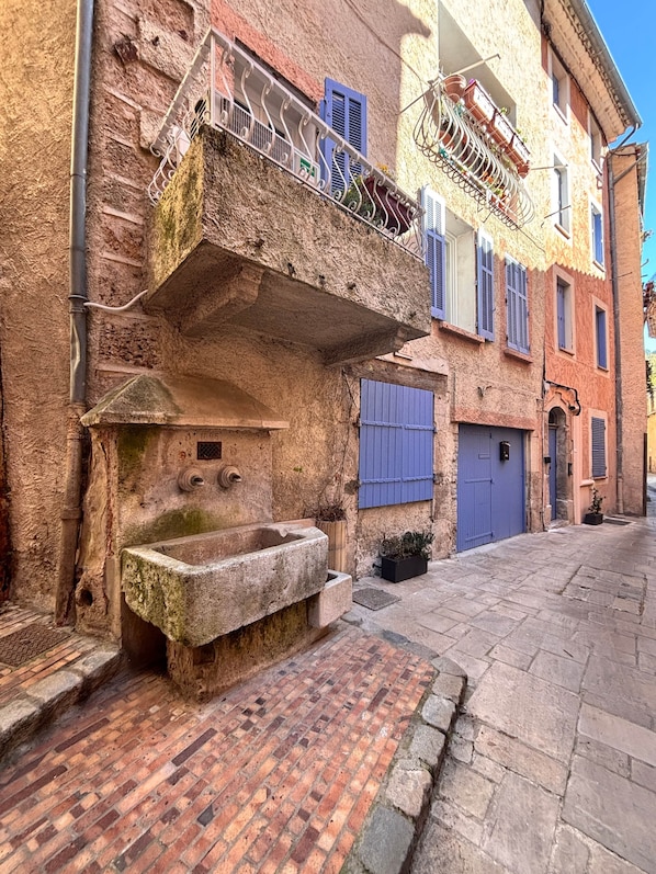 A narrow street with a stone fountain, a metal railing, and a building with a blue door and shutters.