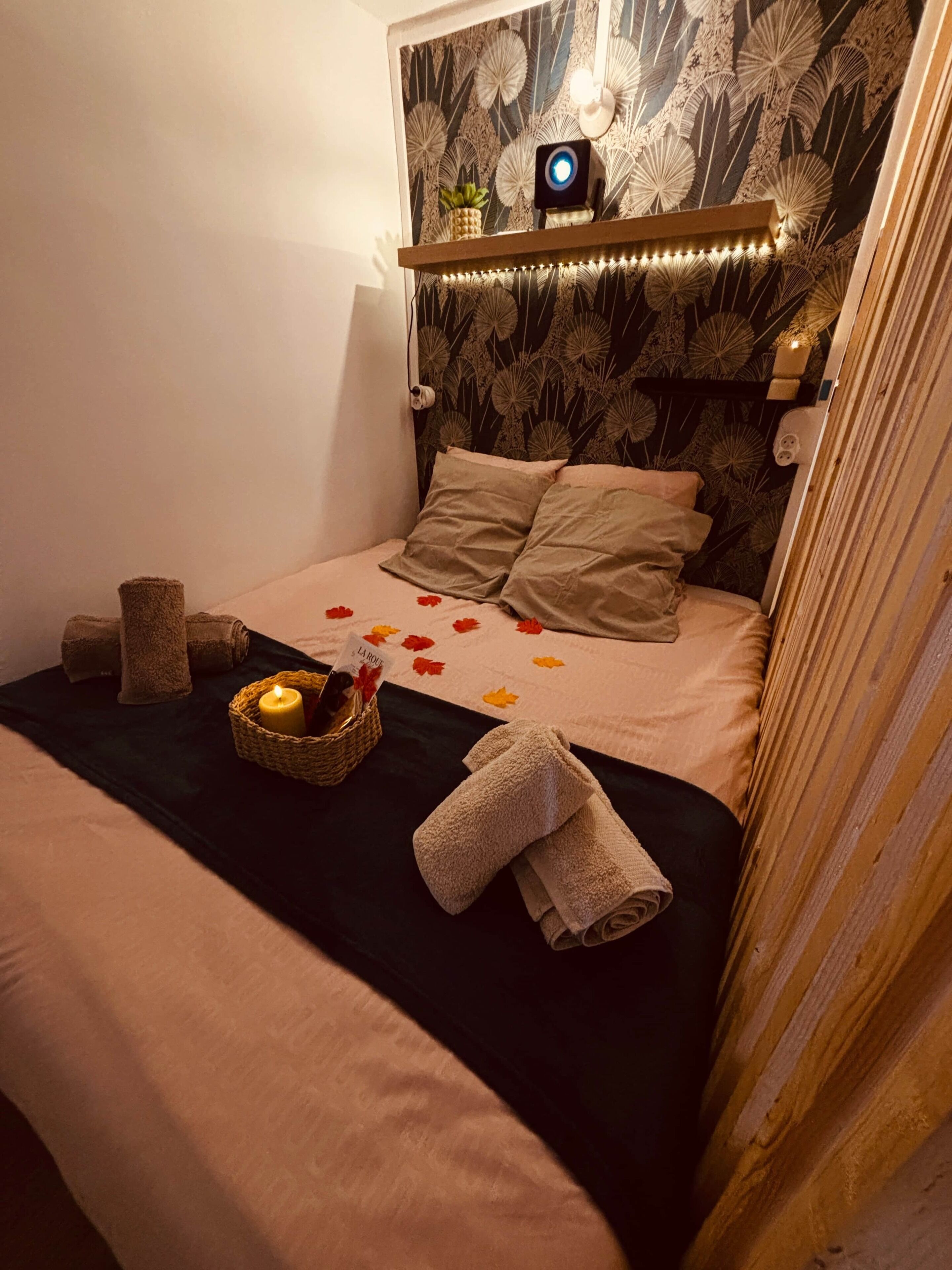 A bedroom with a bed, a wall-mounted light, a small shelf with a plant, and a decorative wall.