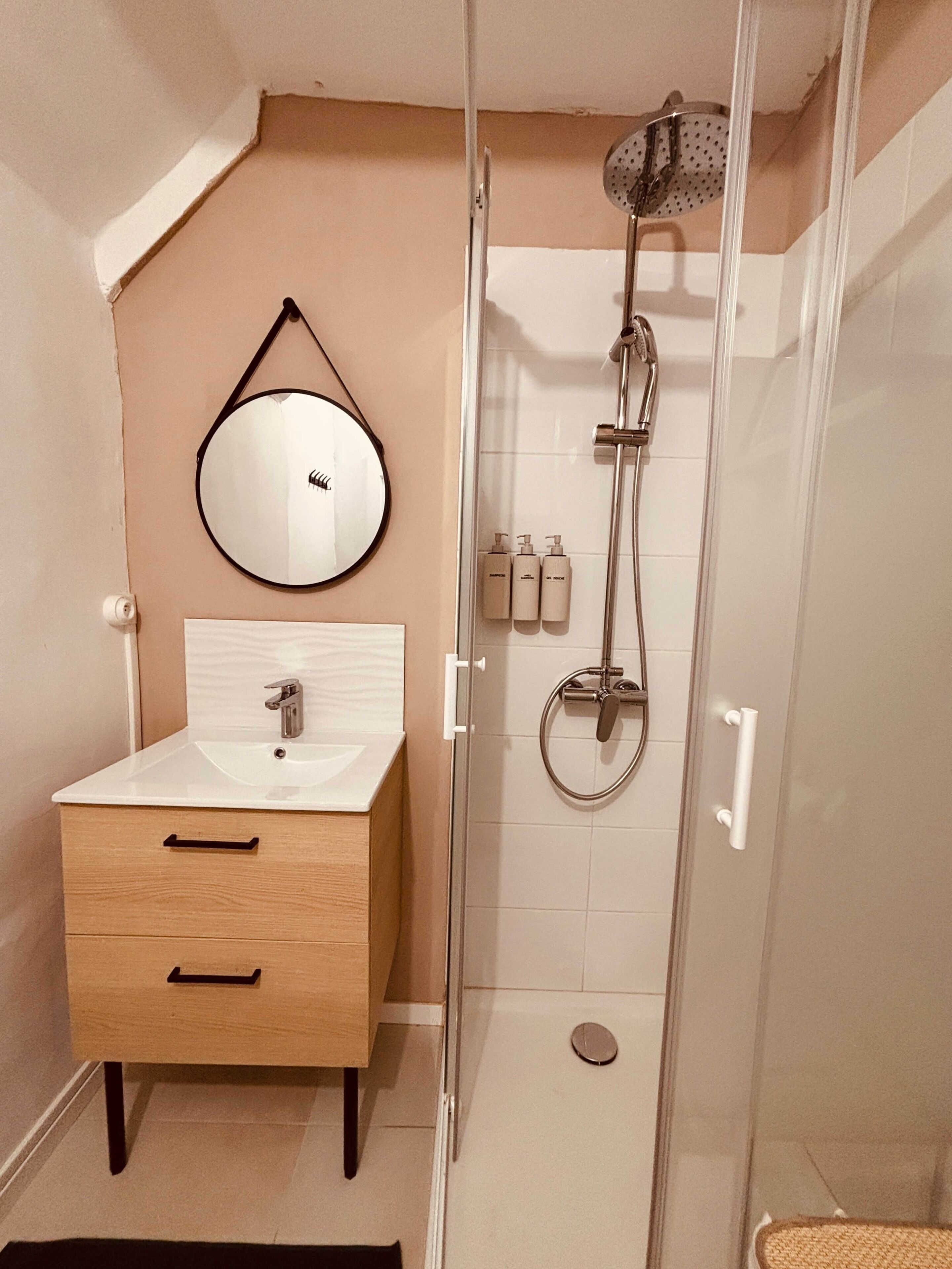 Een badkamer met een houten wastafelmeubel, een ronde spiegel, een douchecabine van glas en een wand betegeld met witte tegels.
