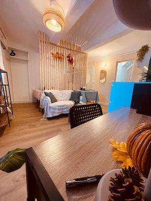 A modern living room with a wooden floor, a sofa, a dining table, and a kitchen area in the background.