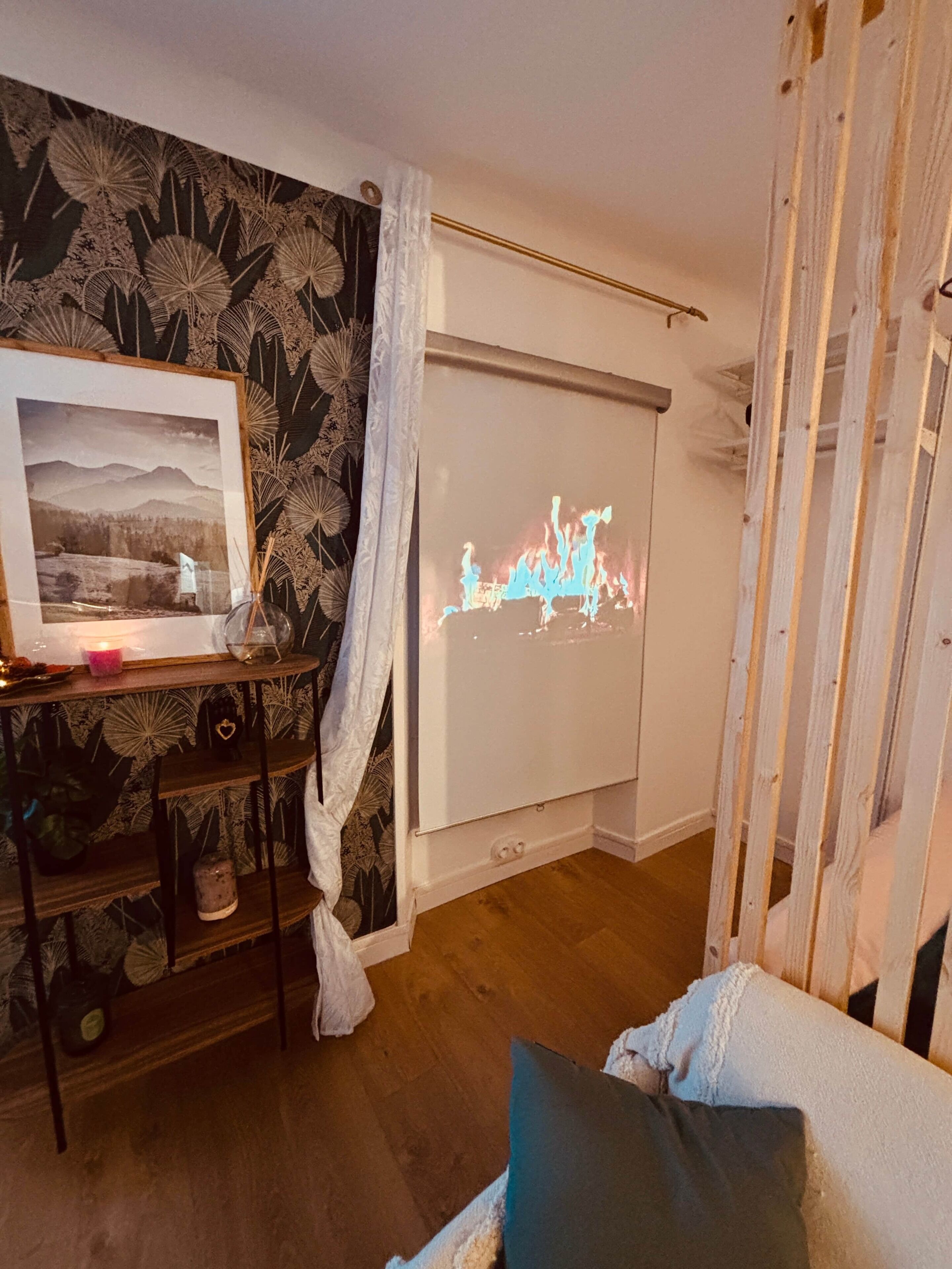 Een kamer met een projectiescherm waarop een vlamanimatie wordt getoond, een ingelijste foto op een plank, en een houten vloer.