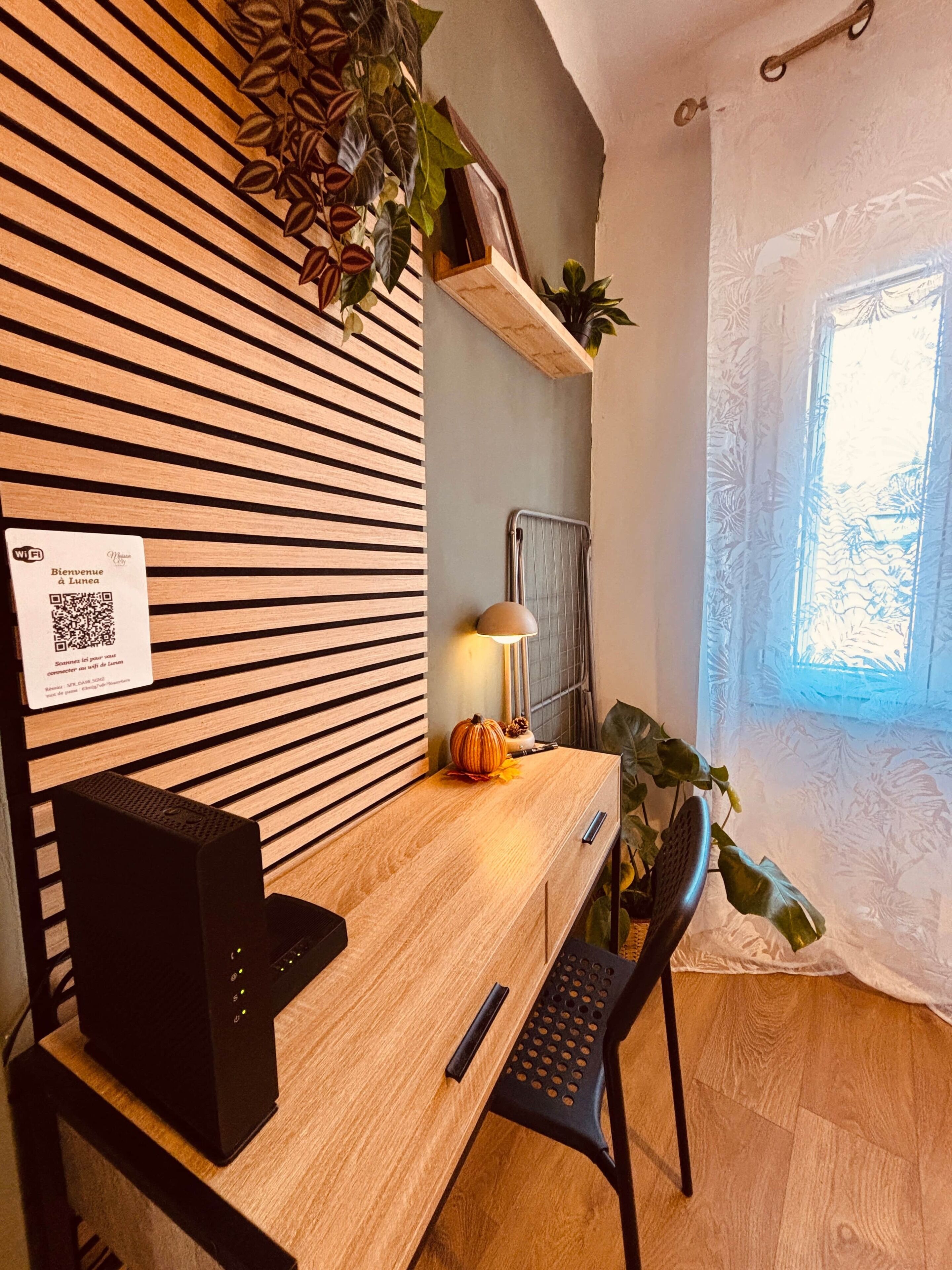 Escritorio de madera con una computadora, una lámpara y una planta. Se ve una ventana con cortina y un radiador.