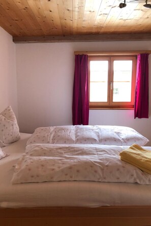 Une chambre avec un plafond en bois, une fenêtre avec des rideaux, un lit avec une literie blanche et une serviette jaune.