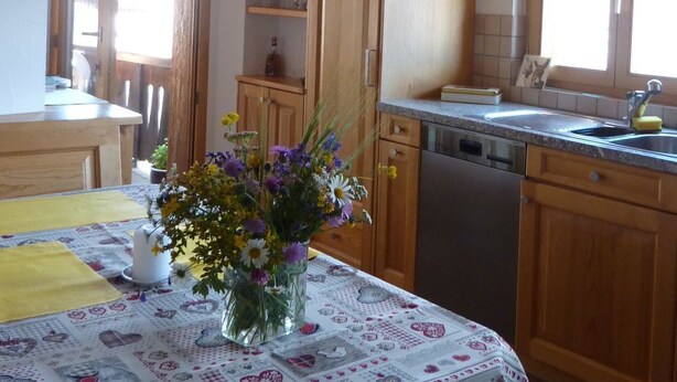 Une cuisine avec des meubles de rangement en bois, une table à manger recouverte d’une nappe à motifs floraux et un luminaire suspendu.