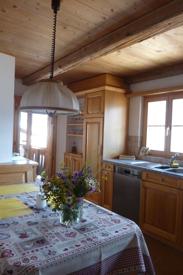 Une cuisine avec des meubles de rangement en bois, une table à manger recouverte d’une nappe à motifs floraux et un luminaire suspendu.