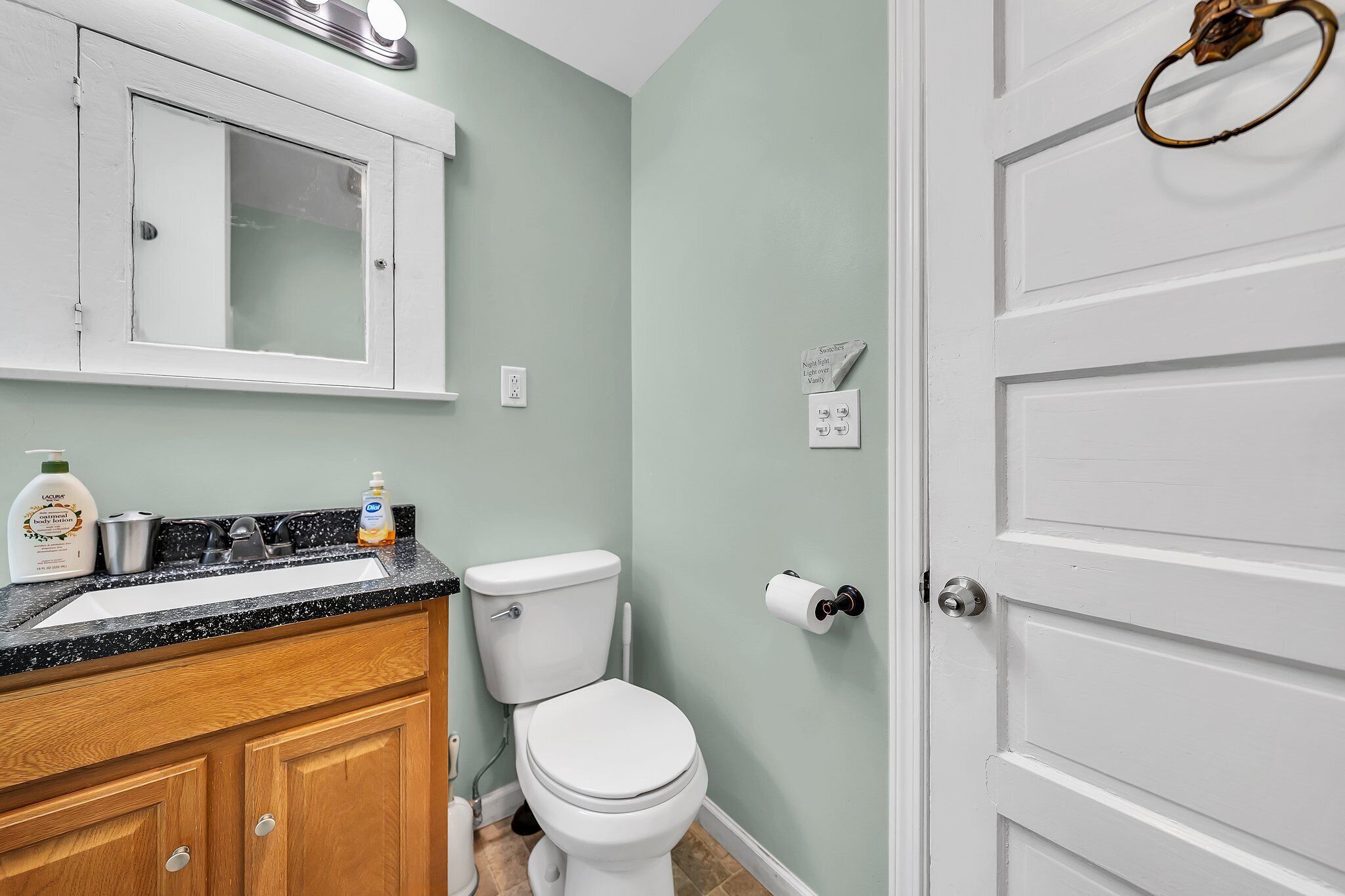 A bathroom with a white toilet, a sink with a black countertop, wooden cabinets, and a white door with a brass handle.