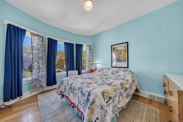 Une chambre à coucher avec un lit, une fenêtre avec des rideaux, une table de chevet, une lampe et un tableau au mur.