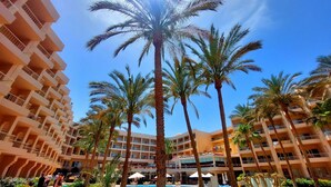 Un hotel de varios pisos con un área de piscina central rodeada de palmeras y un jardín bien cuidado.