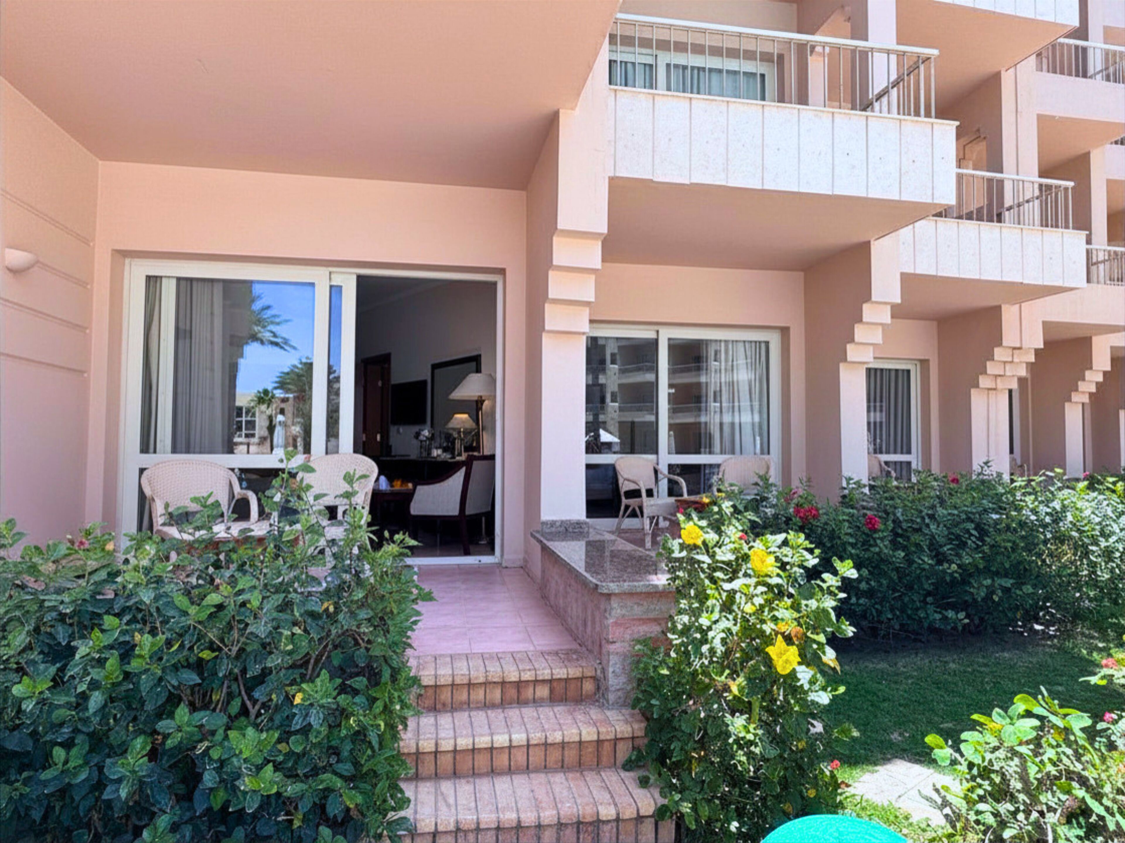 Un hotel con balcón, una escalera que conduce a la entrada y un jardín con arbustos y flores.
