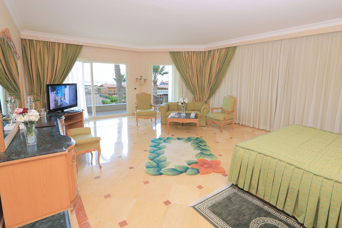 Quarto de hotel com varanda, sofá, mesa de centro e televisão.
