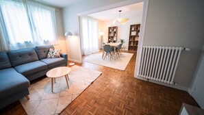 A living room with a sofa, a small round table, a herringbone wooden floor, a radiator, and a dining area with chairs and a table.