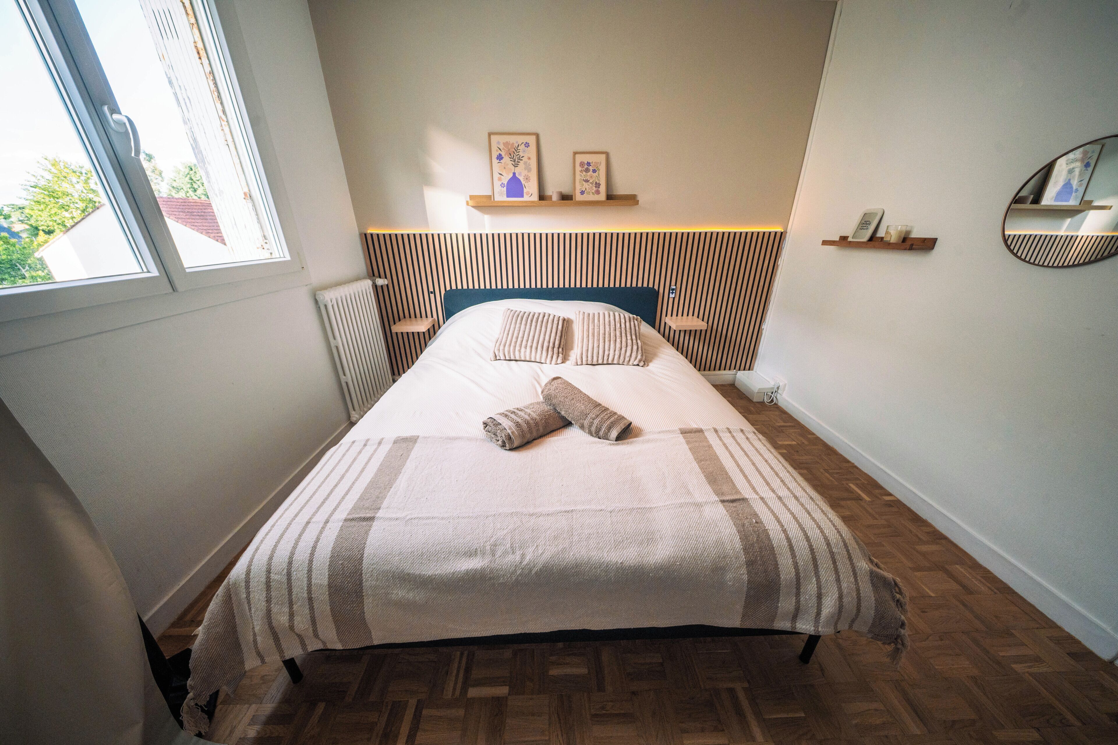Una cama bien tendida con una colcha a rayas y dos almohadas, un cabecero de madera, una ventana con vistas, y una mesita de noche con un teléfono y una taza.