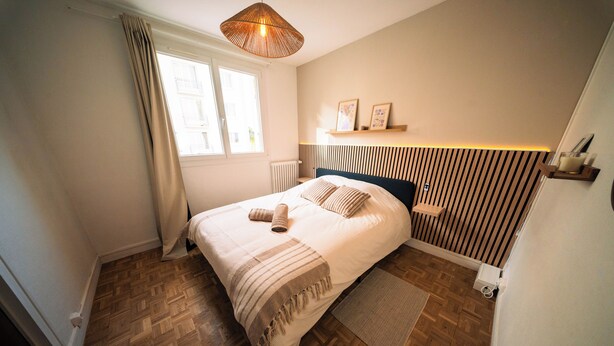 A bedroom with a bed, a window, a radiator, a wooden floor, and a wall-mounted shelf with pictures.