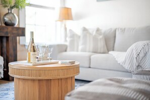 Una mesa redonda de madera con una botella y vasos, un sofá blanco y una lámpara en una sala de estar.