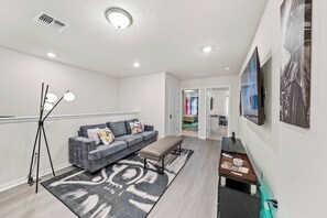 Ein modernes Wohnzimmer mit grauem Sofa, einer Bank, einer Stehlampe und einem an der Wand montierten Flachbildfernseher.
