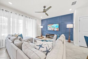 Ein modernes Wohnzimmer mit einer Sofagarnitur, einem an einer blauen Wand montierten Fernseher und einem Deckenventilator.