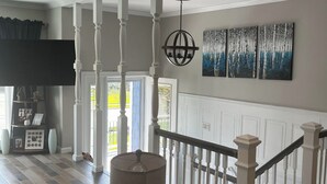 A spacious living room with a staircase, wooden flooring, a large flat-screen TV, and a chandelier.