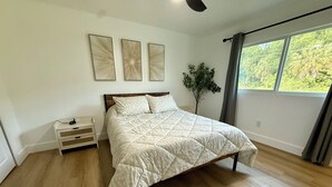 Una camera da letto con un letto, un comodino, una pianta e una finestra vista alberi.