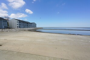 Um edifício com telhado plano e janelas amplas, uma praia arenosa e uma superfície aquática calma.