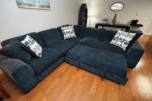 A sectional sofa with geometric patterned pillows, a wooden floor, a bookshelf, a lamp, and a small decorative tree. - Stay Here, Boat There- Walk to Jet + Pool & Beach (Port Clinton)
