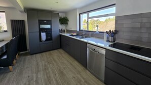 A modern kitchen with a built-in oven, a dishwasher, and a window providing a view of the outdoors. - Hill Head Beach-side Cottage with Hot Tub, Large Garden and Sea Views 5bed slp12 (Fareham)