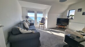 A living room with a sofa, a chair, a TV, and a window with blinds. - Hill Head Beach-side Cottage with Hot Tub, Large Garden and Sea Views 5bed slp12 (Fareham)