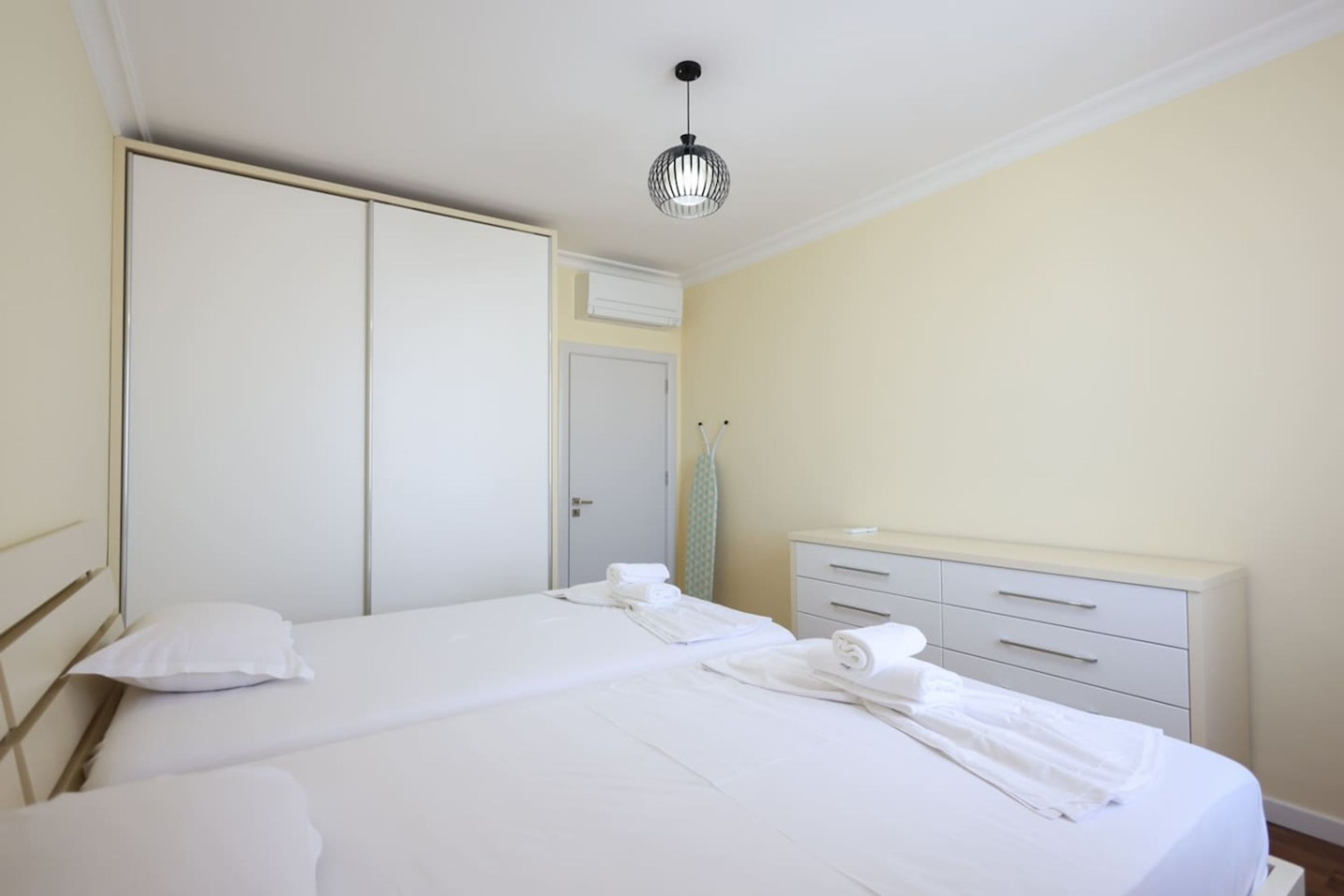 A hotel room with a double bed, a wardrobe, a bedside table with a lamp, and a small desk with drawers.