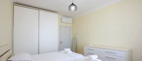 A hotel room with a double bed, a wardrobe, a bedside table with a lamp, and a small desk with drawers.
