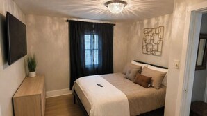 A bedroom with a bed, a TV, a plant, a desk, and a chandelier.