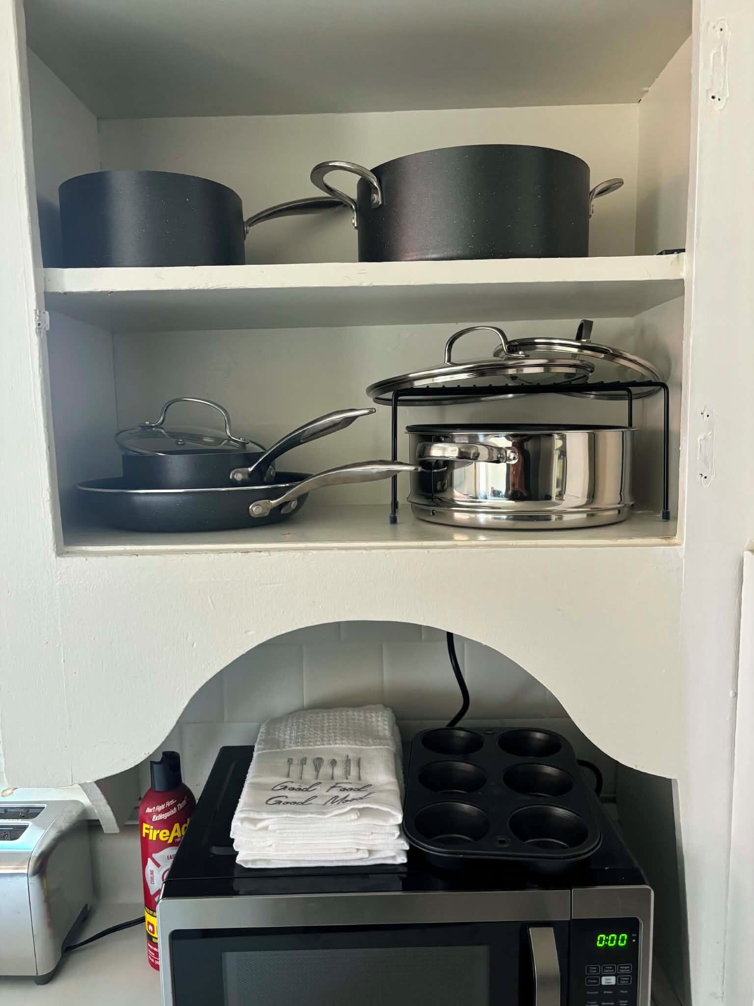 A kitchen with a microwave, a toaster, a fire extinguisher, and pots on shelves.