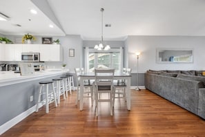 Eine moderne Küche mit weißen Schränken, ein Essbereich mit weißem Tisch und Stühlen, ein graues Sofa und ein großes Fenster.