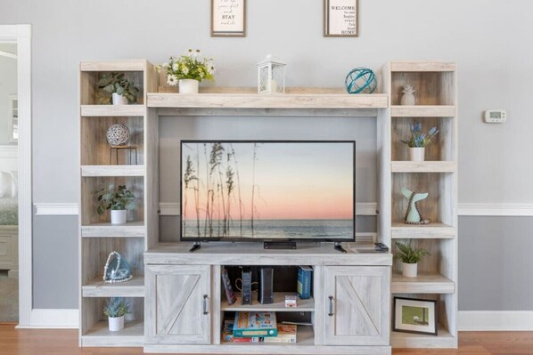 Ein Wohnzimmer mit Fernseher auf einer Holz-Unterhaltungsecke, umgeben von Pflanzen und Dekorationselementen auf Regalen.