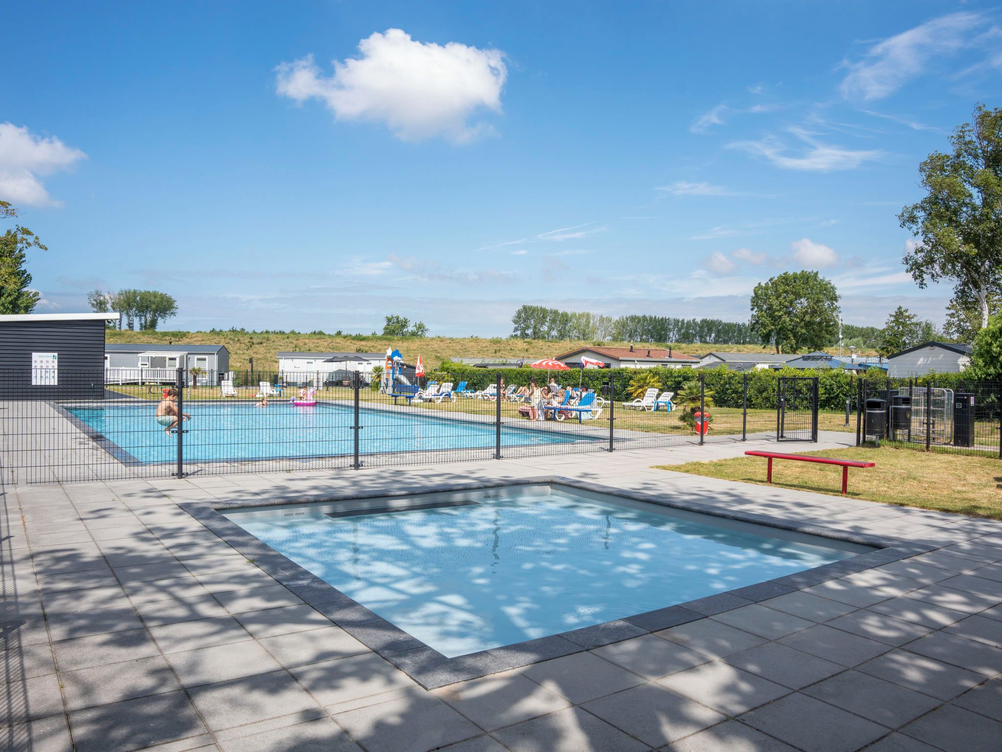 Ein Schwimmbecken mit umzäunter Liegefläche, umgeben von einem angelegten Garten mit Bäumen und wolkenlosem Himmel.
