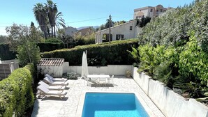 Una piscina rectangular amb terrassa de pedra, envoltada de vegetació i una tanca blanca.