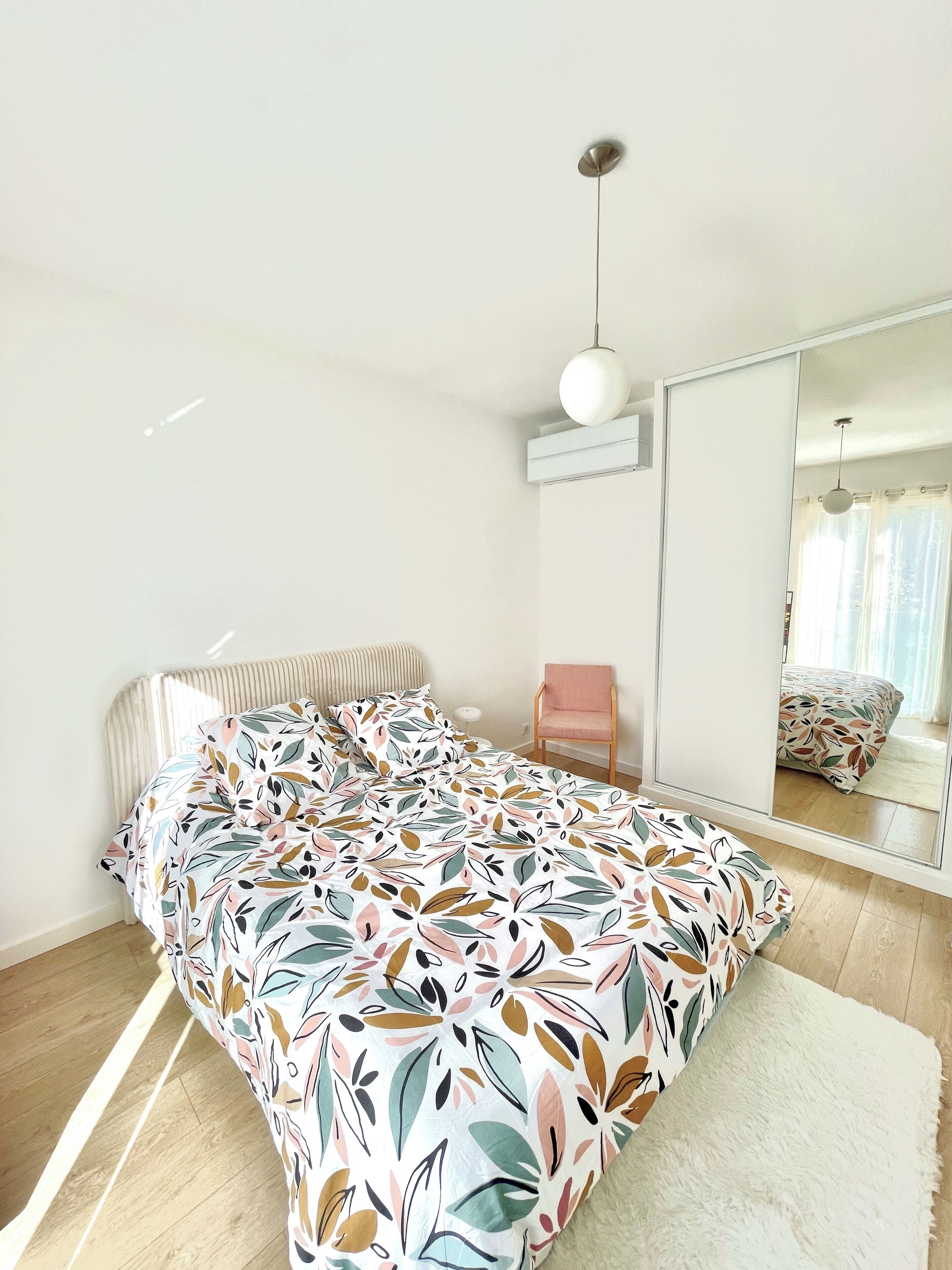 A bedroom with a bed, a chair, a mirror, and a hanging light fixture.