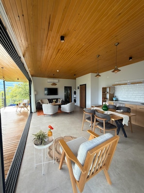 Un vaste espace de vie doté d’un plafond en bois, d’une table à manger avec des chaises, d’un coin cuisine et d’un espace détente avec des canapés et un téléviseur.