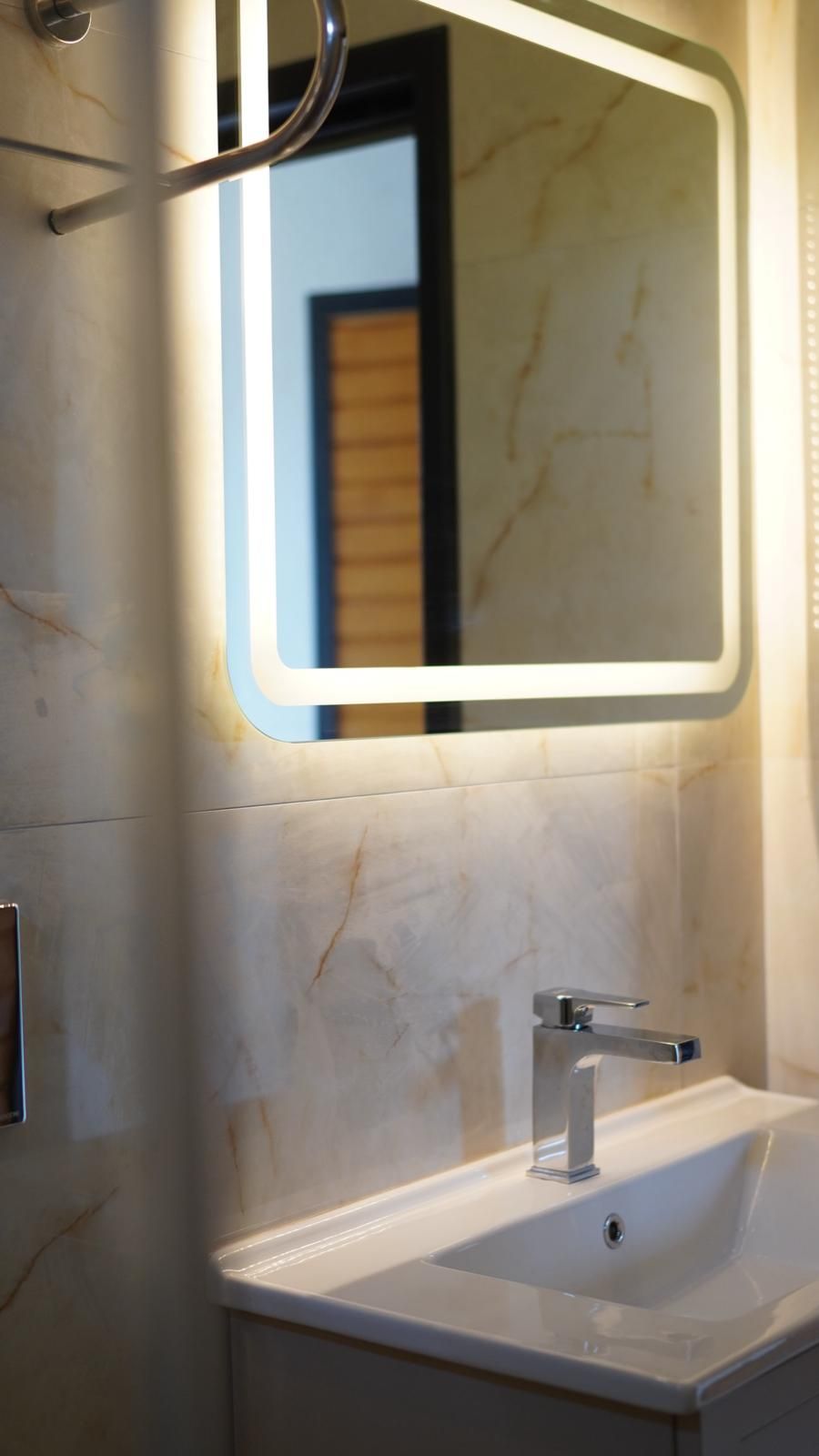 A bathroom with a marble wall, a white sink, and a mirror with a lighted frame.