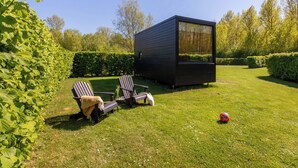 Et moderne udendørs opholdsområde med to Adirondack-stole, et lille bord og en kompakt, sort bygning med et stort vindue.