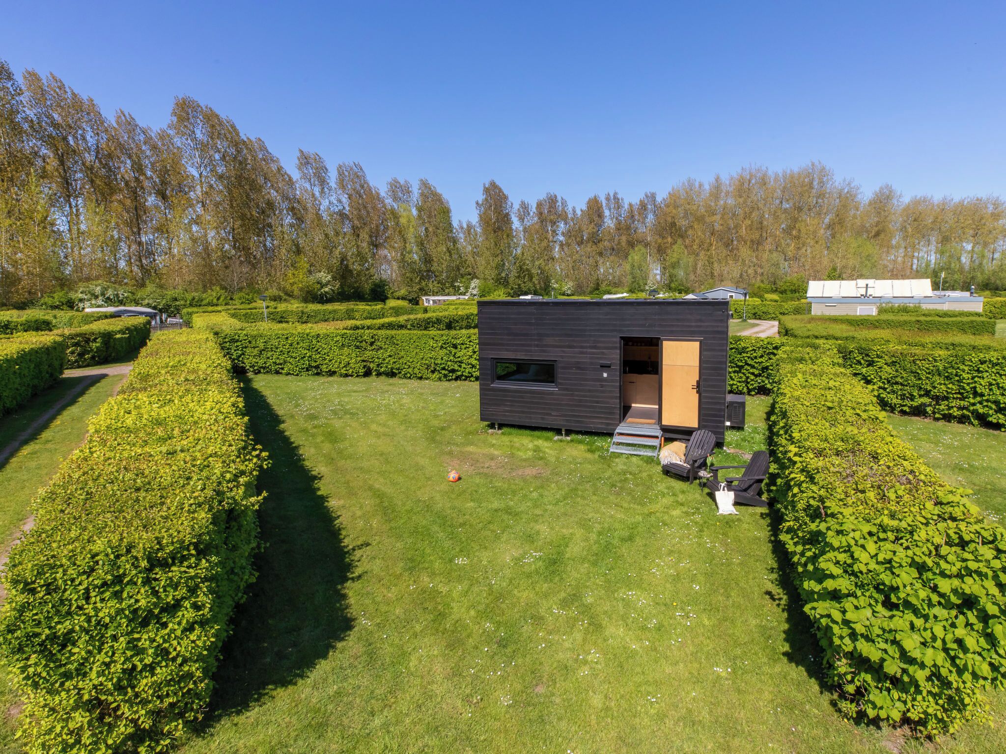 A modern, dark-colored cabin with a small porch, surrounded by well-maintained hedges and a clear sky.